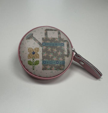 A round, pink coin purse with a floral pattern and a watering can design printed on it.
